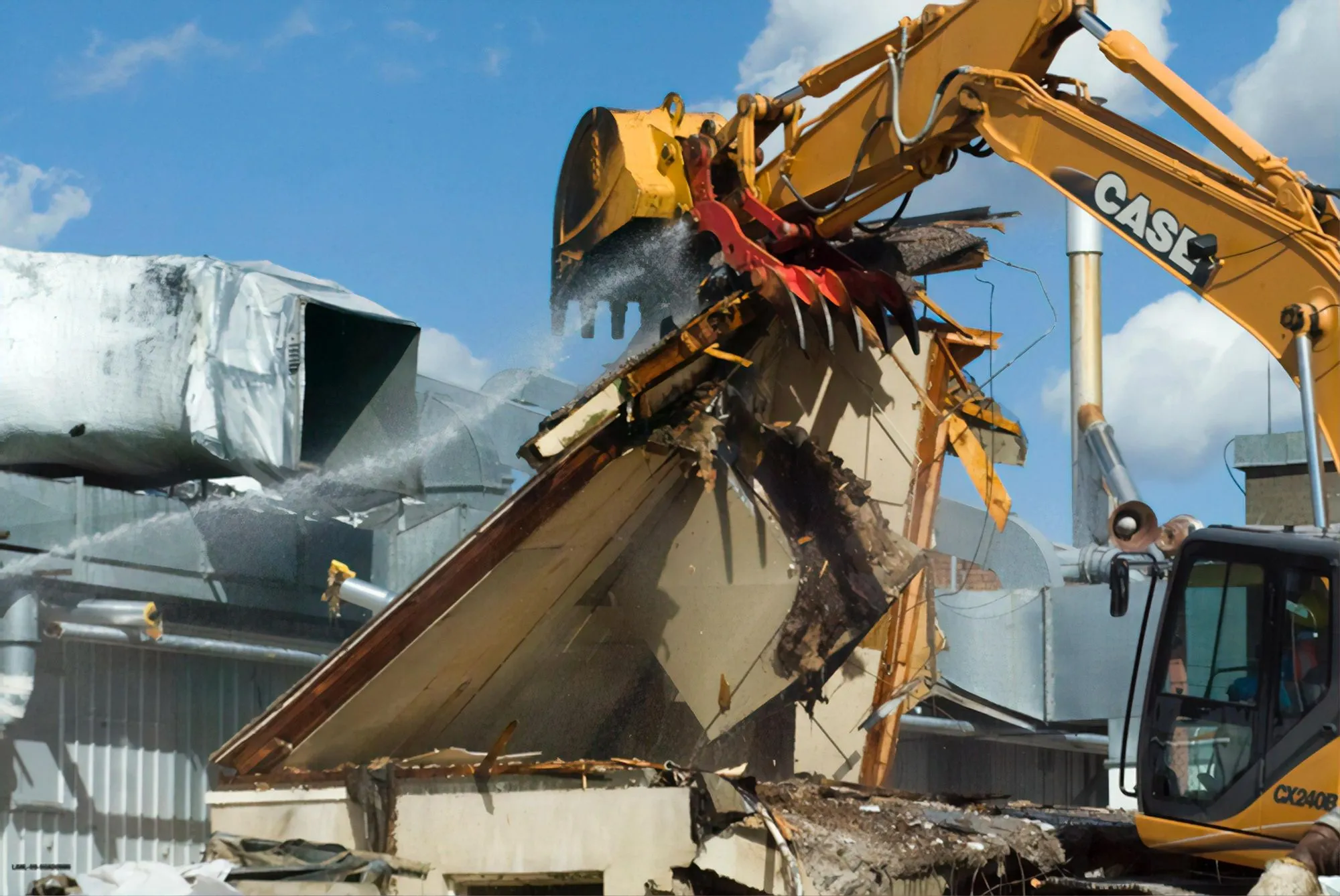 Image of a yellow digger tearing a wall down. There is debris, and the wall is about to fall over. It's set against a blue cloudy sky.